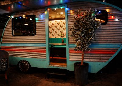 Teal and white vintage camper trailer strung with lights and a potted olive tree