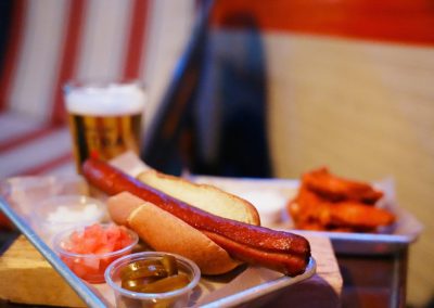 Grilled hot dog on a metal tray with a beer and fries